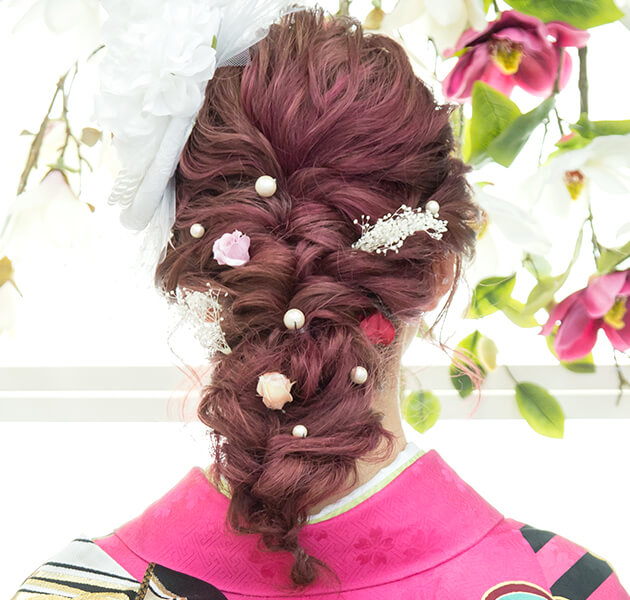 ピンクの髪に白い大きな髪飾りをあしらいパールとピンクのお花をちりばめた編み込みダウンスタイルのモデルの後ろ姿写真