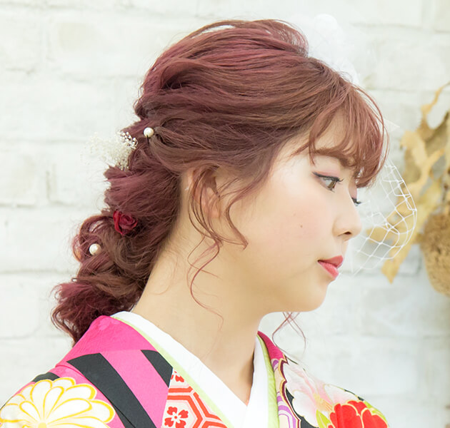 ピンクの髪に白い大きな髪飾りをあしらいパールとピンクのお花をちりばめた編み込みダウンスタイルのモデルの右横顔写真
