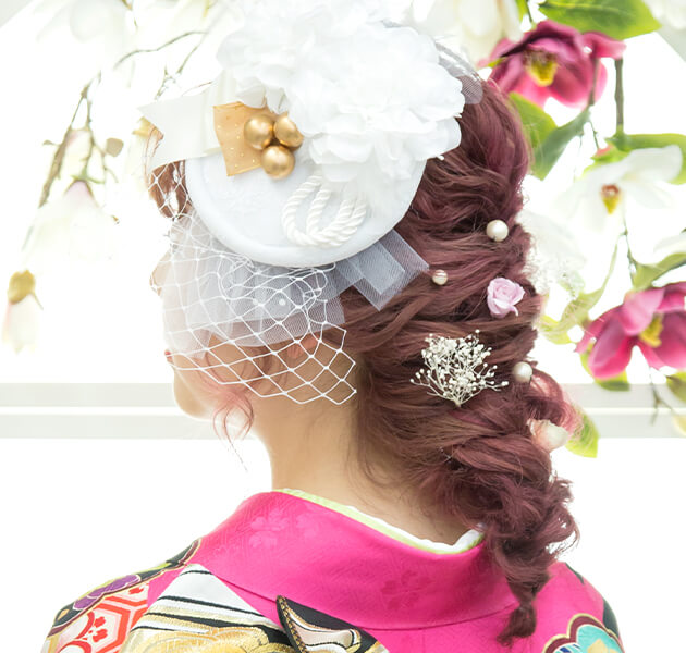 ピンクの髪に白い大きな髪飾りをあしらいパールとピンクのお花をちりばめた編み込みダウンスタイルのモデルの左横顔写真