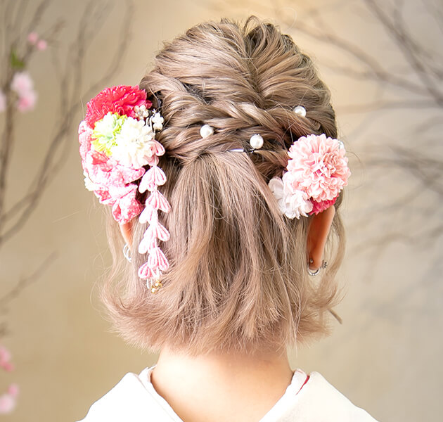 明るいボブヘアーをハーフアップし編み込みとピンクの髪飾りをあしらったダウンスタイルのモデルの後ろ姿