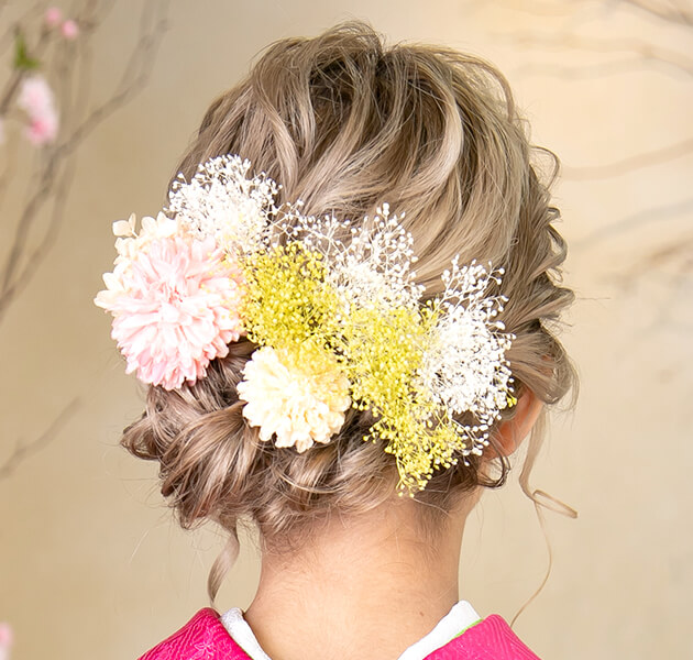 明るい髪色で白、ピンク、黄色の花飾りをしたシニヨンのモデルの後ろ姿写真
