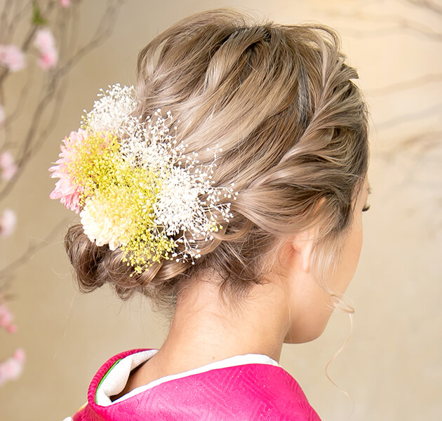 明るい髪色で白、ピンク、黄色の花飾りをしたシニヨンのモデルの斜め右後ろ姿写真