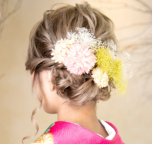 明るい髪色で白、ピンク、黄色の花飾りをしたシニヨンのモデルの斜め左後ろ姿写真